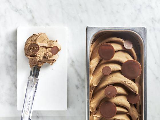 An ice cream scoop containing a generous scoop of milk chocolate ice cream next to a container of gelato garnished with chocolate sauce